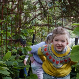 A young girl walking through a wire garden arch followed by other children in a garden filled with vibrant plants.