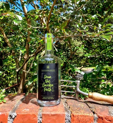 Image a bottle of the 'Down the Garden Path' gin on a brick fence with a tree behind it and a garden fork alongside the bottle. 