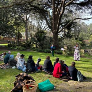 People gathered on a lawn surrounded by trees watching actors perform. 